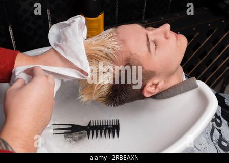 Un coiffeur professionnel essuie les cheveux du client d'un gars avec une serviette blanche au-dessus de l'évier après le lavage, ses cheveux avant une coupe de cheveux dans le salon. Banque D'Images