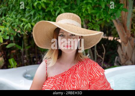 Belle femme caucasienne portant une robe d'été rouge frappante à l'épaule jumelée à un chapeau de paille surdimensionné tout en regardant directement la caméra Banque D'Images