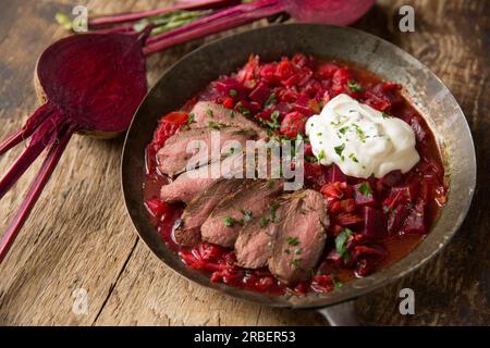 Soupe Borscht maison servie avec des tranches de chevreuil frites à la poêle d'un filet de selle de chevreuil servi avec de la crème fraîche. Affiché dans une casserole sur un cho en bois Banque D'Images