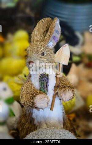 Lapin jouet Bottle Brush avec un regard déterminé sur son visage tenant des fleurs et une bêche de jardin avec des poussins de pâques en arrière-plan - mise au point sélective Banque D'Images