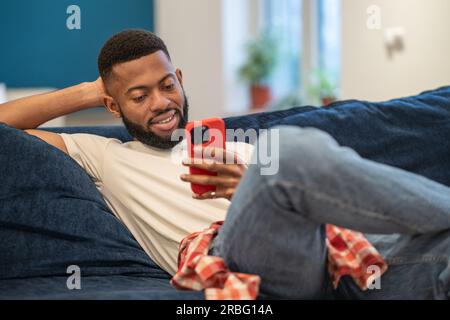 Décontracté souriant africain américain souriant homme assis sur le canapé regarde le smartphone à la maison. Banque D'Images