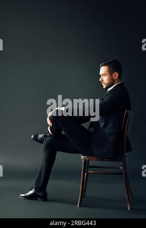 Confiant bel homme en costume noir assis sur la chaise sur fond sombre. Profile view full lenght male portrait Banque D'Images