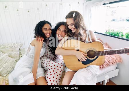 Attrayantes femmes heureuses de différentes ethnies avec guitare s'amusent ensemble à l'intérieur du camping-car. Amitié des femmes et concept de beauté des femmes. Banque D'Images