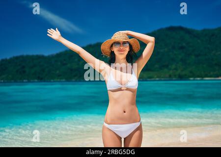 Jeune femme heureuse en bikini blanc et chapeau de paille levés mains levées profiter de ses vacances d'été sur la plage tropicale dans l'île paradisiaque. Détendez-vous, vacances Banque D'Images