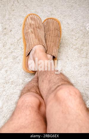 Un homme avec des jambes poilues porte des chaussons chaud et relaxant avec les pieds sur un tapis de laine Banque D'Images