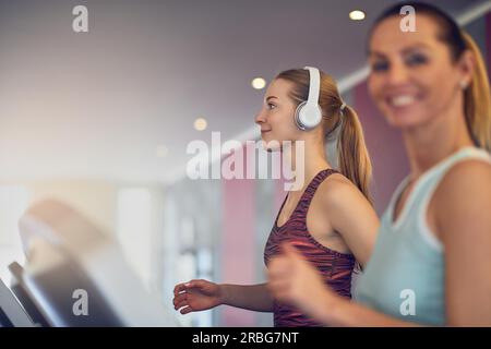 Jolie jeune femme avec des écouteurs sans fil debout sur tapis roulant dans la salle de sport, vue de côté et smiling with copy space Banque D'Images