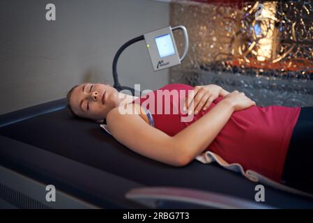 Woman in red shirt portant sur son dos sur table en salle de massage avec écran à côté de sa tête et de verre mur derrière, looking at camera Banque D'Images