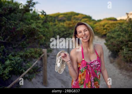 Rire heureuse femme vive en vacances d'été tenant ses chaussures dans sa main alors qu'elle marche sur le sable de la plage le long d'un chemin à travers le Bush Banque D'Images