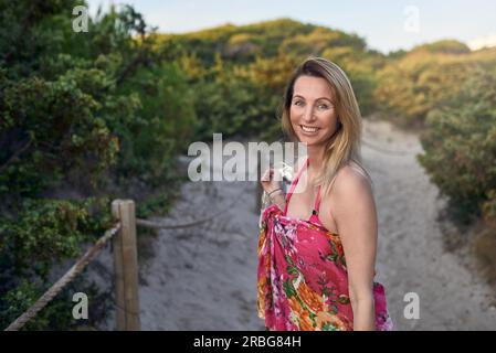 Rire heureuse femme vive en vacances d'été tenant ses chaussures dans sa main alors qu'elle marche sur le sable de la plage le long d'un chemin à travers le Bush Banque D'Images