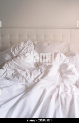 Lit défait avec du linge de lit blanc dans une vue en gros plan d'une couette froissés, des oreillers et une tête de lit rembourrée Banque D'Images
