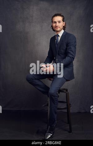 Beau jeune homme aux cheveux longs, portant un costume élégant et une cravate bleue, tenant smartphone dans sa main, regardant l'appareil photo avec un sourire modeste Banque D'Images