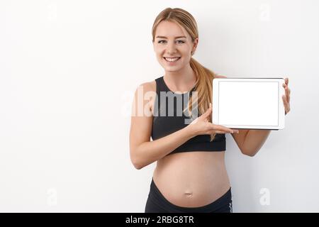 Friendly Smiling young pregnant woman holding up a blank tablet moderne blanc avec écran pour le spectateur sur un fond de studio with copy space Banque D'Images