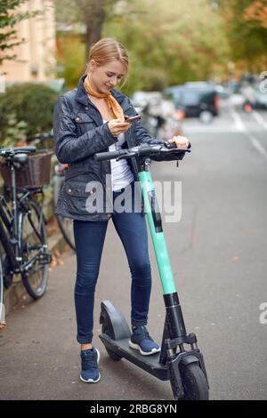 Jeune femme avec trottinette électrique dans la rue de la ville, regardant son téléphone et souriant. Service de location de transport public, moderne Banque D'Images