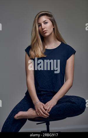 Belle jeune femme en t-shirt bleu, avec les cheveux blonds droits assis avec une jambe sur la chaise et regardant la caméra. Portrait à angle bas avant contre Banque D'Images