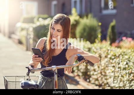 Belle jeune femme avec les cheveux longs dans le portrait avant demi-longueur à l'extérieur dans la ville debout se penchant sur le vélo vintage à l'aide d'un smartphone moderne Banque D'Images