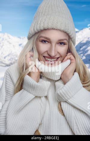 Jolie femme blonde dans un pull col polo en laine chaude et bonnet tricoté debout à l'extérieur se blottissant dans le col avec un large heureux amical Banque D'Images