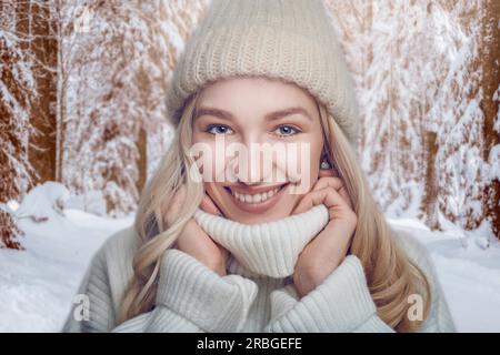Jolie femme blonde dans un pull col polo en laine chaude et bonnet tricoté debout à l'extérieur se blottissant dans le col avec un large heureux amical Banque D'Images