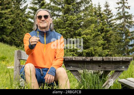 Un homme aux cheveux gris dans des lunettes sombres est assis sur un banc en bois près d'une table et tient une fleur sauvage Banque D'Images