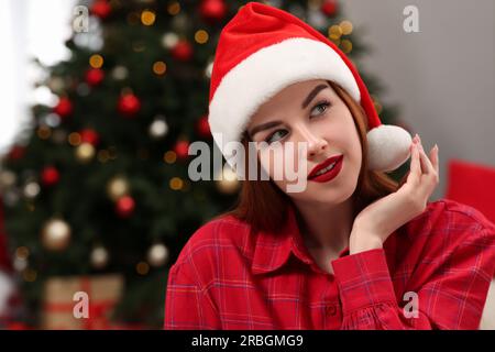 Belle jeune femme dans le chapeau de Santa près de l'arbre de Noël à l'intérieur, espace pour le texte Banque D'Images
