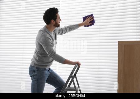 Homme sur l'échelle métallique essuyant les stores à l'intérieur Banque D'Images