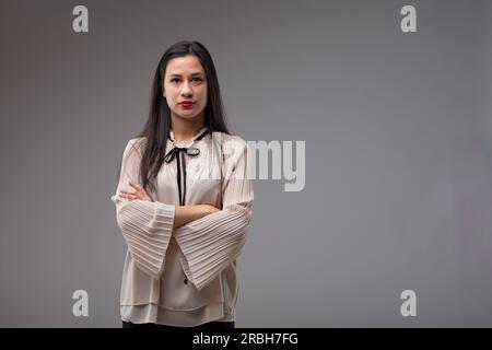 Fière, femme philippine intelligente avec les cheveux longs, les bras croisés. Elle est une patronne confiante dans un chemisier beige avec un ruban noir. Fond gris Banque D'Images