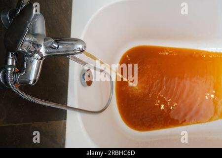 Le robinet de la salle de bain libère un jet de liquide brun. L'eau qui coule du robinet devient orange en raison de la rouille. Banque D'Images