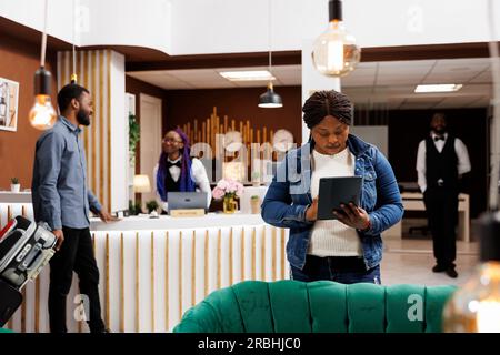 Femme afro-américaine touriste debout dans le hall de l'hôtel avec tablette numérique dans les mains faisant l'auto-enregistrement. Voyageur féminin complétant la procédure d'enregistrement avec un appareil électronique, réservation de chambre en ligne Banque D'Images