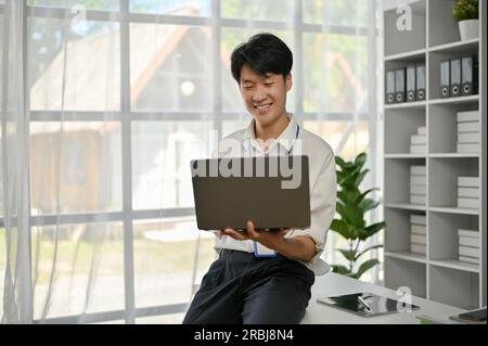 Un heureux et beau jeune homme asiatique employé de bureau ou banquier assis sur une table et utilise son ordinateur portable dans son bureau. Banque D'Images
