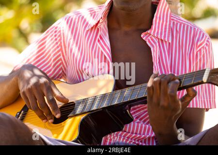 Section médiane de l'homme biracial jouant de la guitare assis sur la plage ensoleillée. Musique, détente, style de vie, été et vacances. Banque D'Images