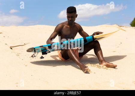 Homme biracial nettoyant planche de surf assis sur la plage ensoleillée. Mode de vie actif, loisirs, sport, surf, été et vacances, non modifié. Banque D'Images