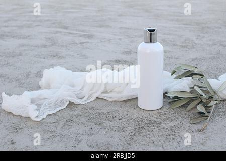 Maquette de bouteille en plastique cosmétique blanche avec branche d'olivier vert et voile de mousseline. Savon liquide naturel, shampooing ou hydratant. Béton grunge gris Banque D'Images