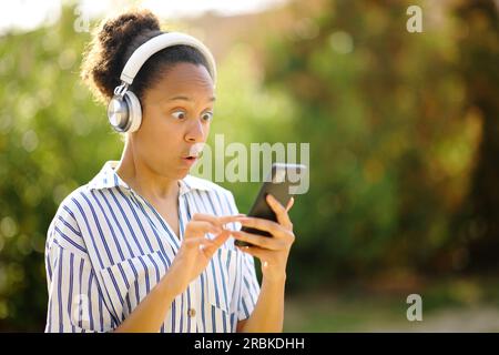 Femme noire surprise portant un casque écoutant de la musique sur le téléphone dans un parc Banque D'Images