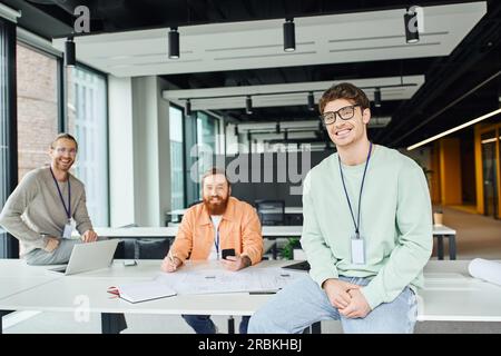 heureux chef d'équipe dans les lunettes regardant la caméra près des architectes souriants travaillant sur le projet de démarrage avec plan, ordinateur portable et téléphone portable dans contempo Banque D'Images