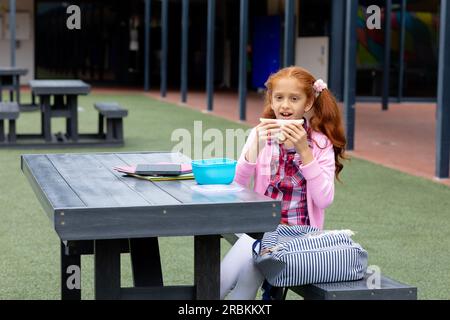 Portrait d'écolière biraciale assise à table dans la cour d'école mangeant un sandwich Banque D'Images