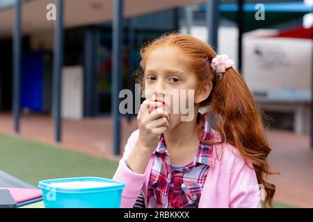 Portrait d'une écolière biraciale assise à table dans la cour d'école mangeant des pommes Banque D'Images