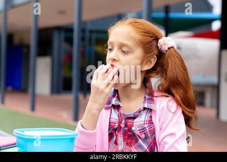 Écolière biraciale assise à table dans la cour d'école mangeant des pommes Banque D'Images