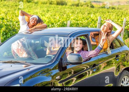 Amis heureux agitant les mains à l'extérieur de la voiture à fenêtre ouverte pendant le voyage sur la route avec les vignobles en arrière-plan Banque D'Images
