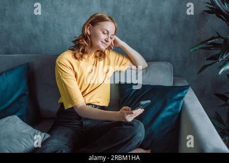 Heureuse jeune fille millénaire vérifiant les médias sociaux tenant smartphone à la maison. Jeune femme blonde souriante à l'aide de l'application de téléphone mobile jouant au jeu, shopping Banque D'Images