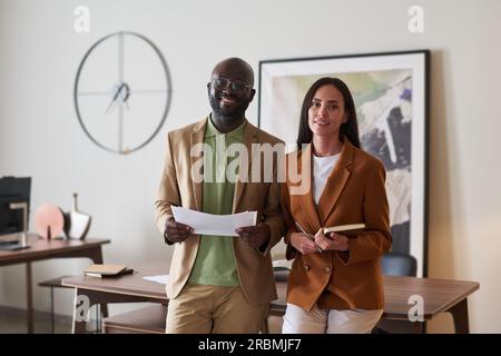 Deux jeunes designers interculturels à succès dans une tenue de luxe tranquille regardant la caméra avec des sourires tout en se tenant près du lieu de travail dans un espace de coworking Banque D'Images