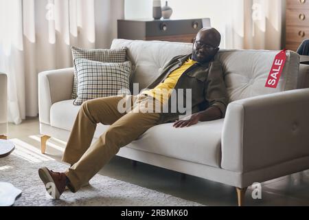 Jeune client masculin afro-américain choisissant un nouveau canapé dans le magasin de meubles ou le département tout en étant assis sur le canapé confortable en velours gris tamisé Banque D'Images