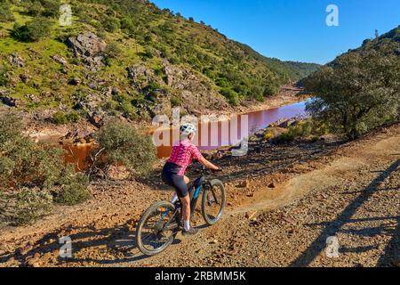 Belle femme avec vélo de montagne électrique sur un tour à vélo le long de la rivière Rio Tinto avec son eau rouge naturelle en Andalousie, Espagne Banque D'Images