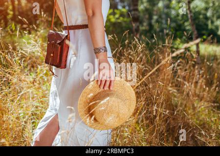Style bohème rustique. Femme élégante portant robe blanche, bijoux, tenant sac à main en cuir et chapeau de paille marchant sur la prairie Banque D'Images