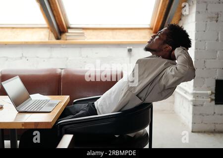Jeune homme d'affaires afro-américain surmené assis dans un fauteuil en cuir devant un ordinateur portable au bureau et étirant les muscles du corps Banque D'Images