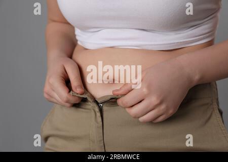 Femme en surpoids essayant de boutonner un pantalon serré sur fond gris, gros plan Banque D'Images