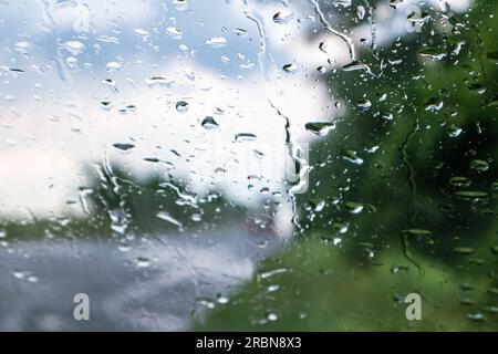 Gouttes de pluie sur la vitre du pare-brise gros plan avec ciel et arbres verts floutés en arrière-plan. Conduite printanière sur la route rurale pluvieuse. Banque D'Images