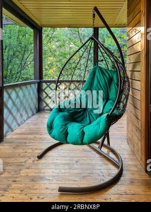 Le concept de détente avec une chaise hamac sur le balcon ou la terrasse. Une chaise balançoire suspendue en osier suspendue sur une chaîne avec un coussin vert .Rattan Banque D'Images