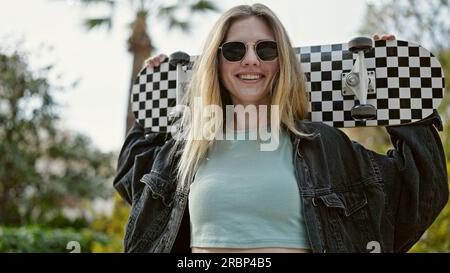 Jeune femme blonde souriante confiante tenant le skate au parc Banque D'Images