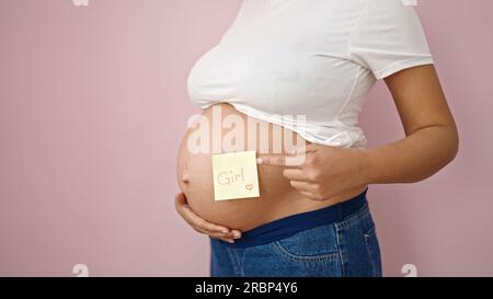 Jeune femme enceinte pointant vers le papier de rappel de fille sur le ventre sur fond rose isolé Banque D'Images