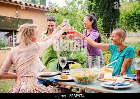 Des personnes mûres enthousiastes levant leurs verres dans un toast joyeux à une fête d'arrière-cour célébrant l'amitié ensemble. Deux hommes d'âge moyen et trois femmes profitant d'un barbecue, clinquant avec du vin. Photo de haute qualité Banque D'Images