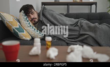 Jeune homme hispanique allongé sur le canapé malade avec des mouchoirs et des pilules sur la table à la maison Banque D'Images